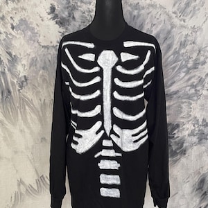 May include: Black long-sleeve shirt with a white skeleton rib cage design on the front. The graphic is hand-painted. The shirt is displayed on a black mannequin against a tie-dye background.