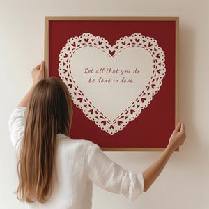 Puede incluir: Una impresión enmarcada con un fondo rojo y un diseño en forma de corazón blanco. El corazón tiene un borde de encaje y el texto "Let all that you do be done in love." El marco es de color marrón claro.