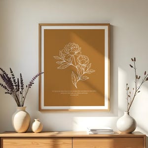 Pode incluir: Uma impressão de arte floral emoldurada com um desenho de linha branca de peónias sobre um fundo mostarda. A impressão inclui uma citação e é exibida acima de uma cômoda de madeira com vasos e plantas decorativas. A sala é iluminada por luz natural.