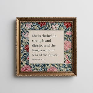 May include: A square framed wall art piece featuring a central beige panel with the black text: She is clothed in strength and dignity, and she laughs without fear of the future. Proverbs 31:25. The border displays a floral pattern in red, pink, and white.