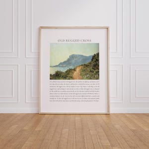 May include: A framed print titled "OLD RUGGED CROSS" features a landscape painting of a coastal path leading to a distant mountain. The artwork is in a wooden frame and hangs on a white wall. The print includes text below the image.