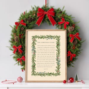 May include: A framed print of "The Christmas Story" text, surrounded by a green and blue garland. A Christmas wreath with red bows and pinecones hangs behind the print. Red and white ornaments and a small Christmas tree are in front.