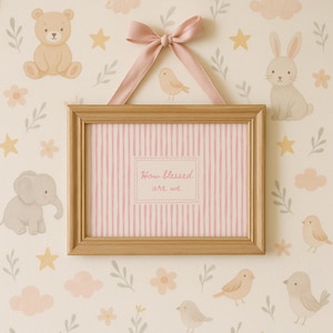 May include: A framed picture with a pink and white striped background and the text "How blessed are we." The frame is made of light brown wood and is suspended by a pink ribbon. The background features illustrations of a bear, bunny, elephant, birds, stars, clouds, and flowers.