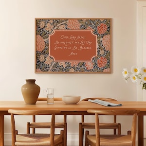 Op de afbeelding: Een ingelijste print met een bloemenpatroonrand en een centraal tekstvak. De tekst luidt: "Come, Lord Jesus. Be our guest and let thy gifts to us be blessed. Amen." De print wordt weergegeven boven een houten eettafel.