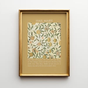 May include: A framed wall art print featuring a gold border and a central botanical pattern of green leaves and tan fruits on a cream background. The text HAIL MARY appears at the top, with the full Hail Mary prayer printed in white at the bottom.