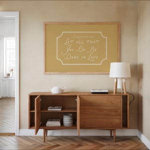 Puede incluir: Un aparador de madera con puertas abiertas, que muestra libros y un jarrón decorativo. Arriba, un cartel enmarcado de color mostaza dice: "Que todo lo que hagas sea con amor". Una lámpara blanca con base de madera está sobre el aparador.