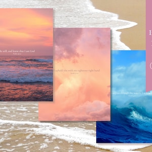 Three 18x14-inch poster prints with inspirational bible verses set against coastal scenes. The first print features a pink and orange sunset over the ocean with the verse 'Be still, and know that I am God.' The second print features a pink and orange cloudy sky with the verse 'I will Uphold you with my righteous right hand.' The third print features a blue ocean wave with the verse 'when you pass through these waters, I will be with you.'