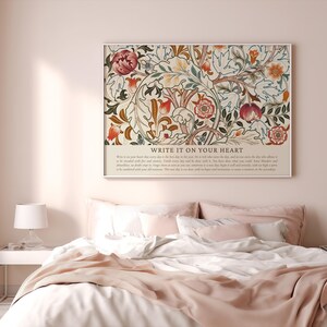 May include: A framed floral art print with the text "WRITE IT ON YOUR HEART" hangs above a bed. The artwork features a cream background with red, orange, and green floral patterns. The bed has white linens and a pink blanket.