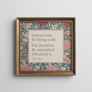 May include: A square framed wall art piece featuring a beige central panel with the black text: Instructions for living a life: Pay attention. Be astonished. Tell about it. Mary Oliver. The border displays a dark blue floral pattern.