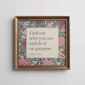 May include: A square framed wall art piece featuring a central beige panel with the black text: Find out who you are and do it on purpose. Dolly Parton. The panel is surrounded by a dark blue floral pattern with red and pink flower motifs.