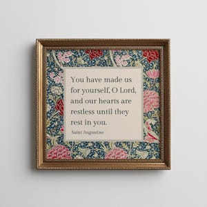 Könnte beinhalten: Ein quadratisches gerahmtes Wandbild mit einem zentralen beigen Feld und dem schwarzen Text: You have made us for yourself, O Lord, and our hearts are restless until they rest in you. Saint Augustine. Der Rand zeigt ein florales Muster in Dunkelblau.