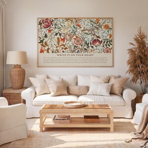 May include: A framed floral art print with the text "WRITE IT ON YOUR HEART" above a white sofa with beige pillows. A wooden coffee table and a woven lamp are in the foreground. The artwork features red, orange, and green floral patterns.