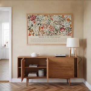 May include: A wooden sideboard with open doors, displaying shelves and a decorative vase. Above, a framed floral print with the text "WRITE IT ON YOUR HEART" hangs on the wall. A white lamp with a wooden base sits on the sideboard.