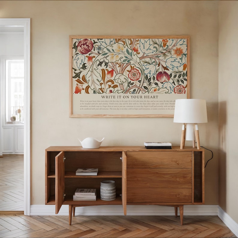 May include: A wooden sideboard with open doors, displaying shelves and a decorative vase. Above, a framed floral print with the text "WRITE IT ON YOUR HEART" hangs on the wall. A white lamp with a wooden base sits on the sideboard.