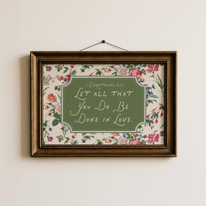 Op de afbeelding: Een ingelijste print met een bloemenontwerp en de tekst "Let all that you do be done in love." De lijst is donkerbruin. De achtergrond heeft een crèmekleur met een bloemenpatroon van roze, rode en blauwe bloemen.