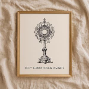 May include: A framed print featuring a black and white detailed illustration of a monstrance centered on a cream background. Below the illustration, the text reads: BODY, BLOOD, SOUL & DIVINITY. The frame is a thin, metallic gold color.