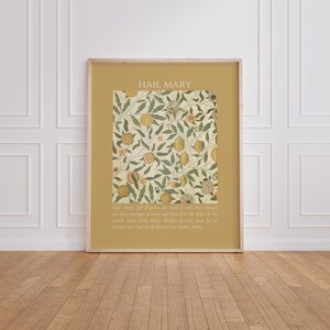 May include: A rectangular wall art print with a mustard yellow border featuring the text HAIL MARY at the top and the full prayer text at the bottom. The center displays a botanical pattern of green leaves and orange fruits on a cream background.