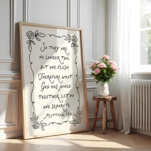May include: A framed print with the text "So they are no longer two, but one flesh. Therefore what God has joined together, let no one separate. Matthew 19:6." The frame is light-coloured wood. The print is displayed next to a vase of pink roses.