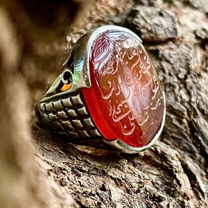May include: A silver ring with a red carnelian stone featuring Arabic calligraphy. The stone is oval shaped and set in a silver band with intricate details.