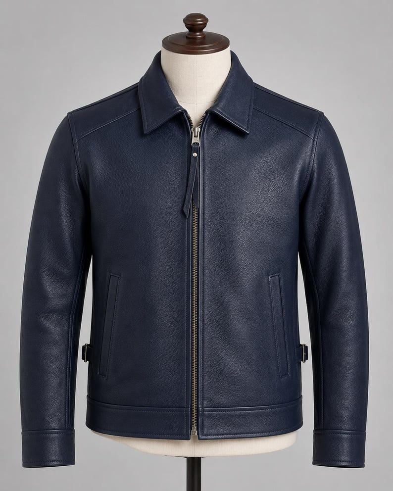 May include: A navy blue jacket with a textured finish, featuring a classic collar, a central metal zipper closure with a long pull tab, two vertical side pockets, and adjustable side straps at the hem, displayed on a neutral-toned mannequin.