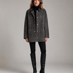 May include: A charcoal gray suede jacket featuring a pointed collar, long sleeves, and a front button closure with silver-toned hardware. The jacket includes two large patch pockets on the front, paired with a black turtleneck, black leggings, and black knee-high boots.