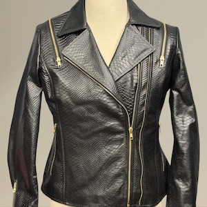 Może przedstawiać: Czarna kurtka ze sztucznej skóry o fakturze skóry węża. Kurtka posiada kołnierz z klapami, liczne kieszenie zapinane na zamek i złote zamki. Kurtka jest prezentowana na manekinie.