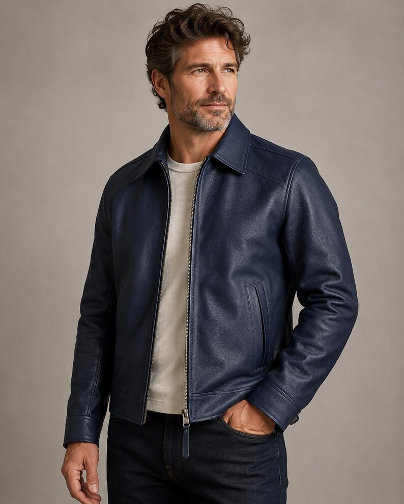 May include: A navy blue leather jacket featuring a classic collar, a full-length silver metal zipper closure, and two vertical side pockets. The jacket is worn over a plain white crew neck t-shirt and dark denim jeans, showcasing a smooth texture.