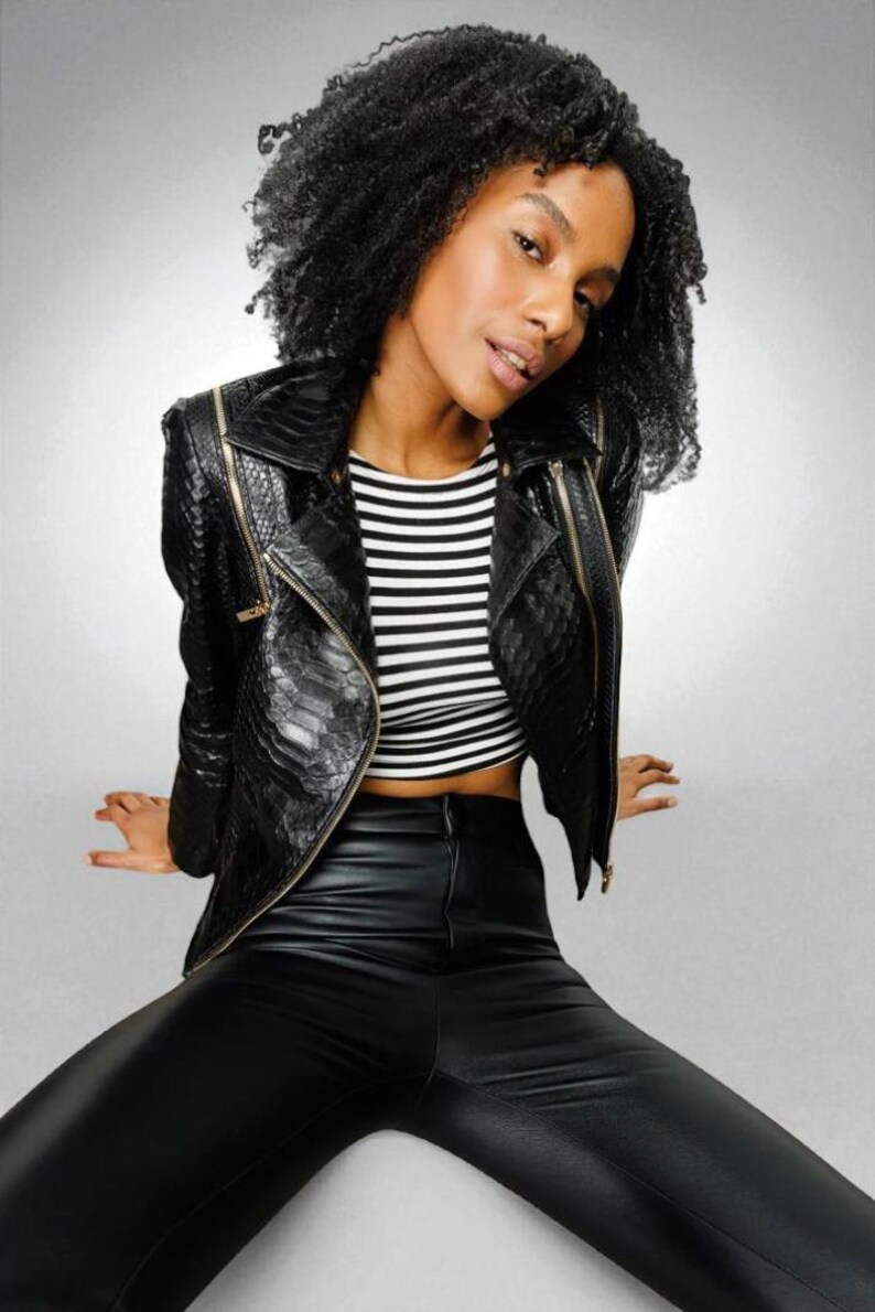 May include: A black leather jacket with gold zippers, paired with black leather pants and a black and white striped crop top. The jacket has a textured pattern. The model is sitting with legs spread.