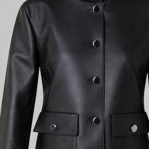 May include: A black leather jacket with a round collar and long sleeves. The jacket features four large, shiny silver buttons down the front and two flap pockets with matching buttons. The brand name "A-K-R-I-S-" is visible on the upper back.