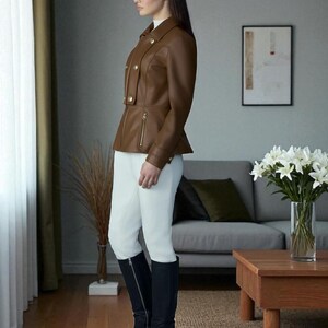 May include: A brown leather jacket with gold buttons and a zipper, paired with white pants and black knee-high boots. The jacket has a tailored fit. The boots have a block heel. The outfit is suitable for equestrian or formal wear.