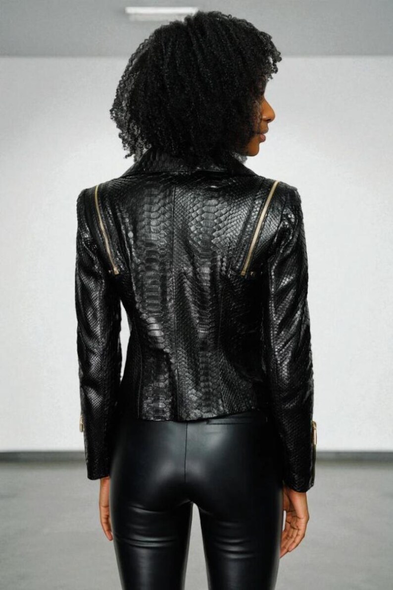 May include: A black faux leather jacket with a snakeskin texture and gold zippers. The jacket has a tailored fit and a classic biker style. The model is wearing black leather pants. The background is a plain white wall.