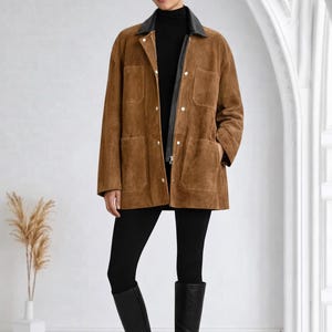 May include: A brown suede jacket with a black leather collar and four front pockets. The jacket is worn over a black turtleneck and paired with black leggings and knee-high black boots.