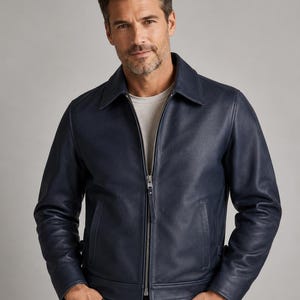 May include: A navy blue leather jacket featuring a classic collar, a full-length silver-toned metal zipper closure, long sleeves with cuffs, and two vertical side pockets. The jacket is worn over a plain light gray crew neck t-shirt.