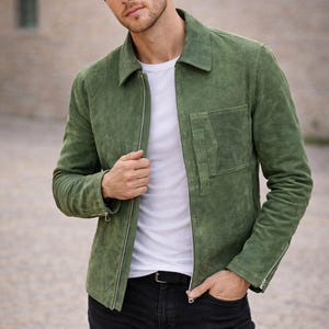 May include: A green suede jacket with a zipper closure, worn over a white t-shirt and paired with black jeans. The jacket has a collared neckline and a pocket on the chest.