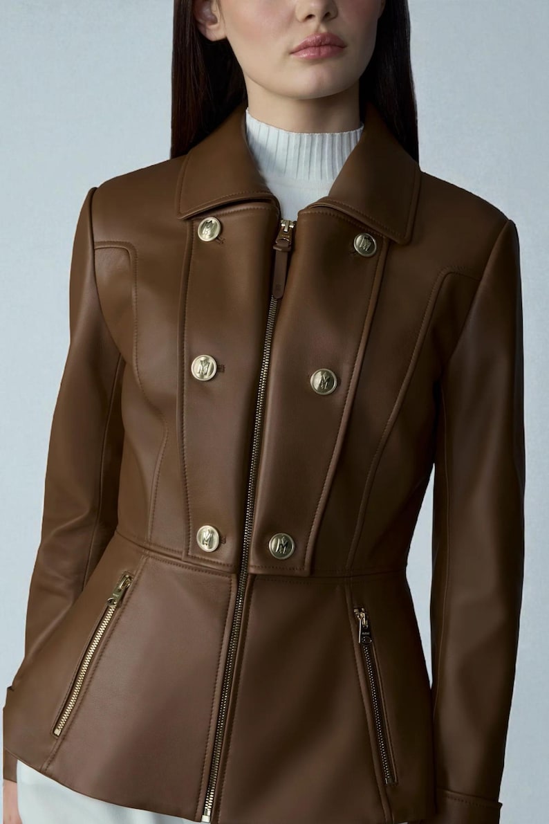 May include: A brown leather jacket with a tailored fit, featuring a collar, gold-colored buttons, and a zipper closure. The jacket has two zippered pockets and a fitted waist. The model is wearing a white turtleneck underneath.