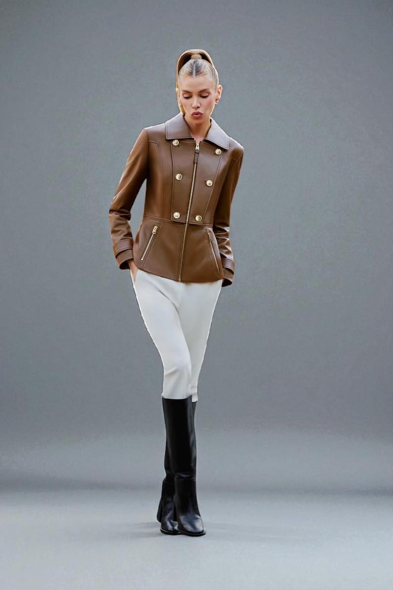 May include: A brown leather jacket with gold buttons and zippers, paired with white pants and black boots. The jacket has a double-breasted design and a tailored fit, creating a stylish and sophisticated look. The outfit is suitable for various occasions.