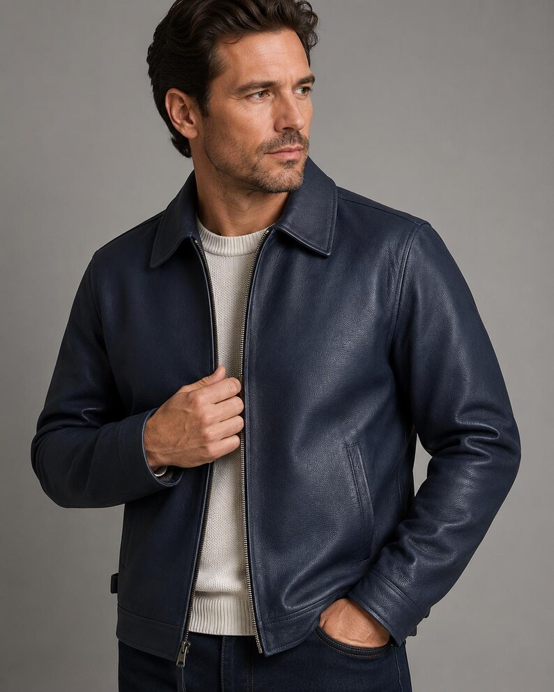 May include: A navy blue leather jacket featuring a classic collar, a full-length silver-toned front zipper, and two side pockets. The jacket is worn over a white crew-neck sweater, showcasing a smooth texture and a structured, tailored fit.