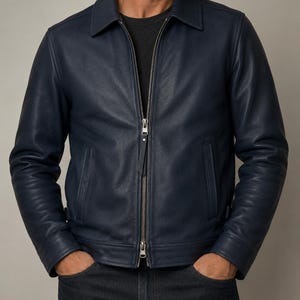 May include: A navy blue leather jacket featuring a classic collar, long sleeves, and a central silver-toned metal zipper closure. The jacket includes two vertical side pockets and a smooth, matte finish, worn over a plain black t-shirt and dark jeans.