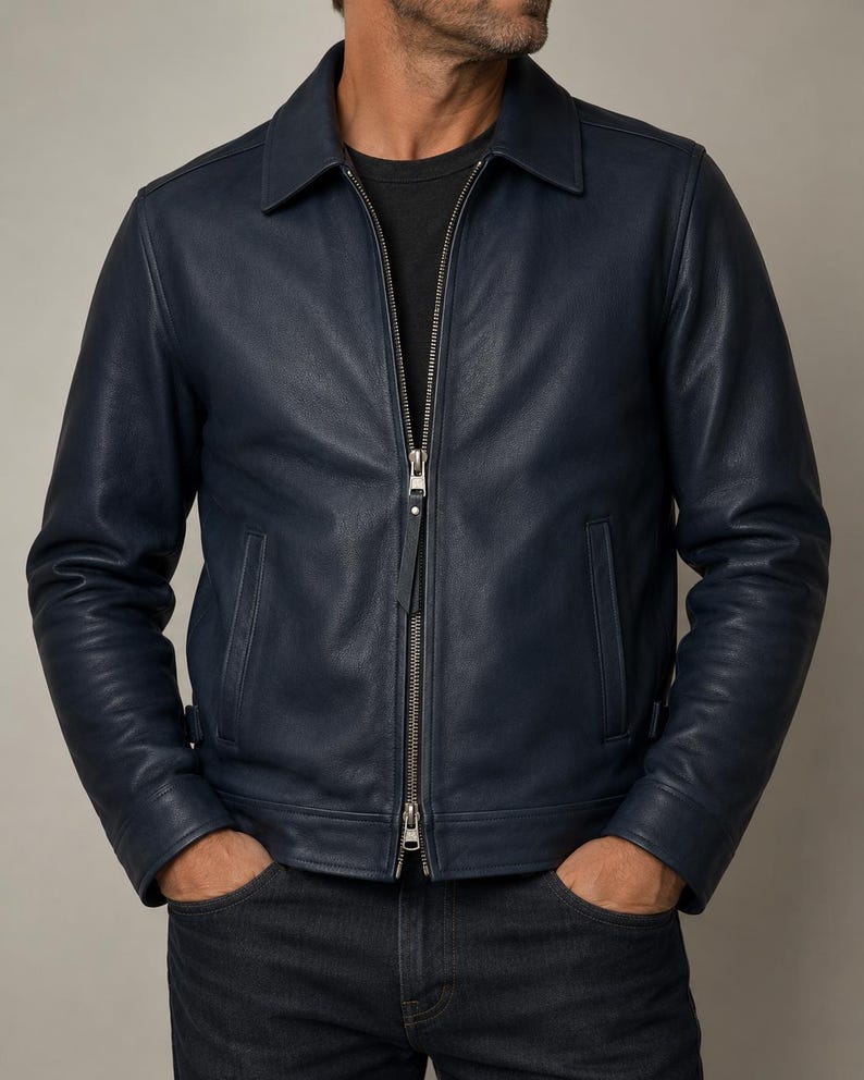 May include: A navy blue leather jacket featuring a classic collar, long sleeves, and a central silver-toned metal zipper closure. The jacket includes two vertical side pockets and a smooth, matte finish, worn over a plain black t-shirt and dark jeans.