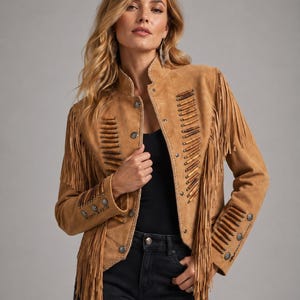 May include: A tan suede jacket with fringe detailing. The jacket features decorative wooden beads and silver-toned buttons. The jacket has a stand-up collar and is worn over a black top and black jeans. The jacket is a western-inspired style.