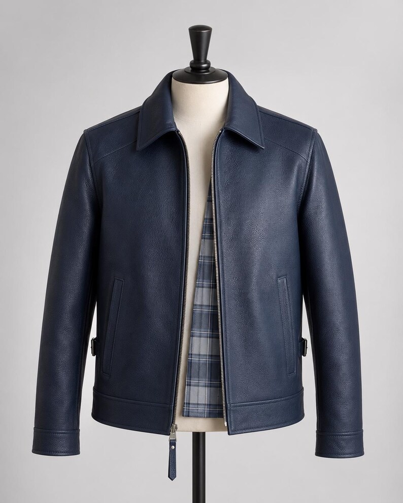 May include: A navy blue jacket with a textured finish, featuring a classic collar, a full-length silver metal zipper, and two vertical side pockets. The interior lining displays a grey and blue plaid pattern, and the cuffs have a button detail.