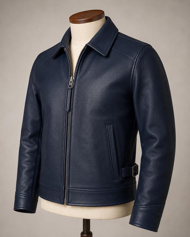 May include: A navy blue leather jacket featuring a classic collar, a full-length front metal zipper closure, long sleeves with cuffs, and a side waist adjustment strap with a metal buckle. The jacket has two vertical welt pockets at the front.