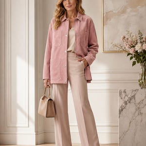 May include: A woman's outfit featuring a light pink suede jacket with front pockets, a cream-colored top, and matching wide-leg pants. A beige handbag and pointed-toe heels complete the ensemble. The background includes a painting and a vase of flowers.