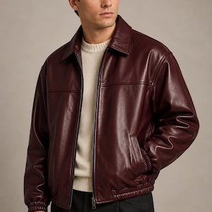 May include: A deep burgundy leather jacket featuring a classic collar, a full-length front zipper, and ribbed elastic cuffs and hem. The jacket is worn over a cream-colored ribbed knit sweater and dark charcoal trousers.