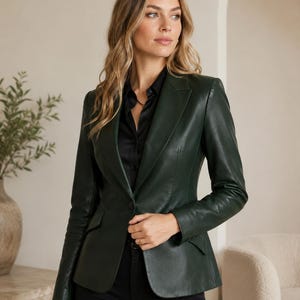 May include: A dark forest green leather blazer featuring a single-button closure, notched lapels, and two front flap pockets. The jacket is worn over a black collared shirt and black trousers, showcasing a structured fit with long sleeves.