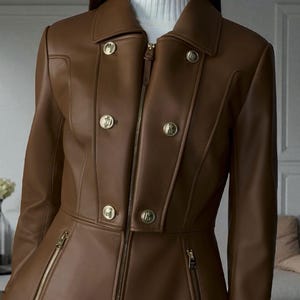 May include: A brown leather jacket with a tailored fit. The jacket features a notched collar, gold-tone buttons, and zippered pockets. The model is wearing a white turtleneck underneath. The jacket is a stylish outerwear piece.