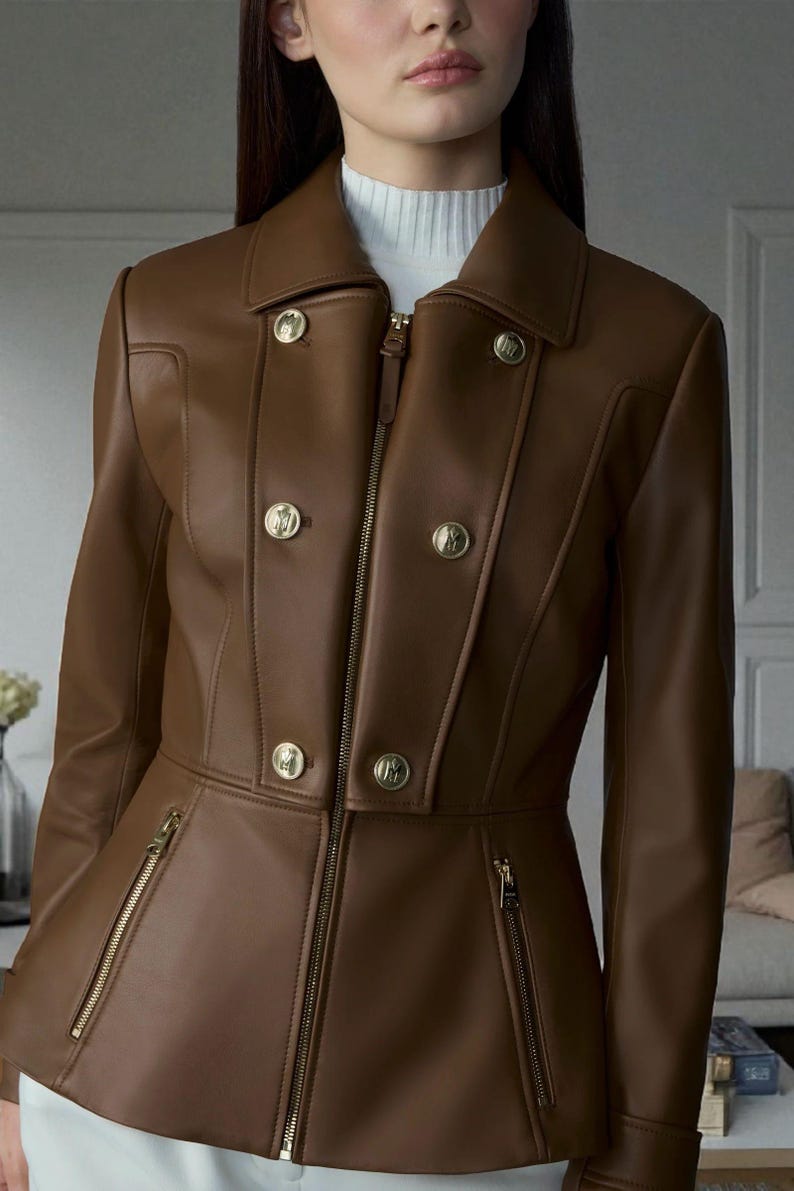 May include: A brown leather jacket with a tailored fit. The jacket features a notched collar, gold-tone buttons, and zippered pockets. The model is wearing a white turtleneck underneath. The jacket is a stylish outerwear piece.