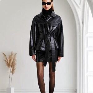 May include: A black leather jacket with a matching mini skirt. The jacket has a belted waist and two front pockets. The outfit is paired with black tights and heels. The jacket and skirt have a crocodile skin texture.