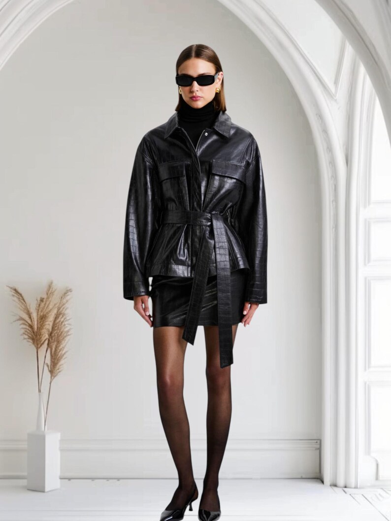 May include: A black leather jacket with a matching mini skirt. The jacket has a belted waist and two front pockets. The outfit is paired with black tights and heels. The jacket and skirt have a crocodile skin texture.
