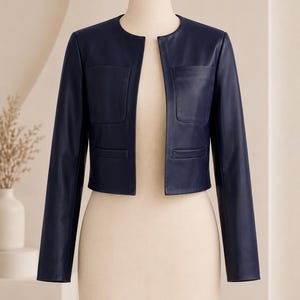 May include: A navy blue cropped jacket with a collarless round neckline and long sleeves. The jacket features two square chest pockets and two lower welt pockets, with a smooth finish and an open front design displayed on a mannequin.